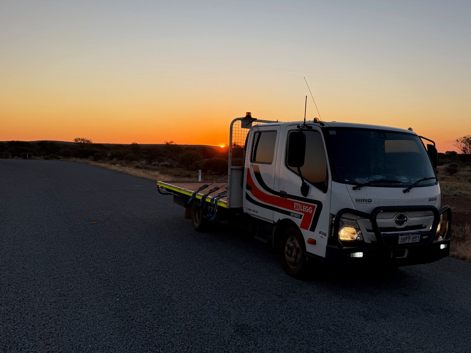 JM Legg Hotshots Hino crew-cab truck with an alloy tray, parked on an open road at sunset with its headlights on